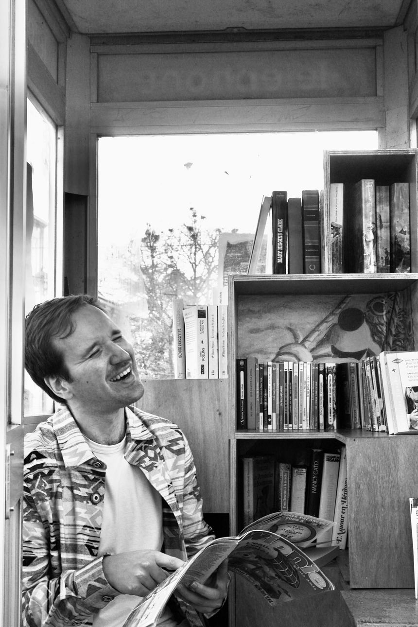 Titelive, en noir et blanc, rit en lisant un magazine dans une cabine téléphonique transformée en bibliothèque